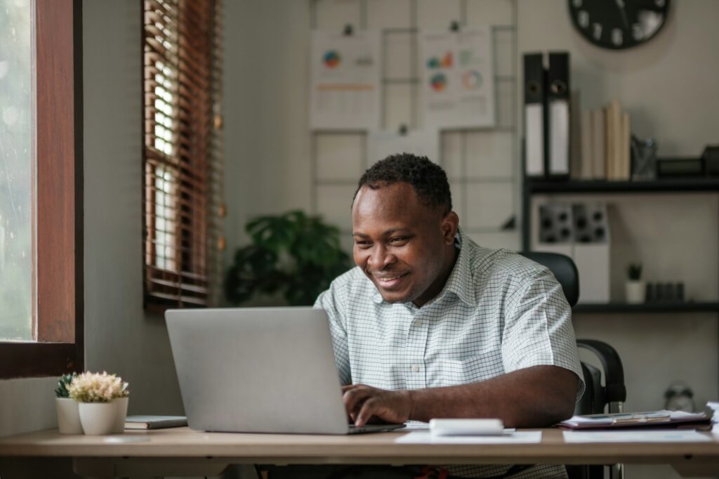 Smiling black man using laptop at home in living room. Happy mature businessman send email and